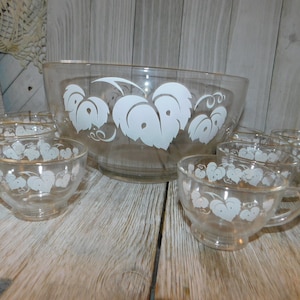 May include: A vintage glass punch bowl and cups set with a white grape leaf design. The bowl and cups are clear glass with a gold rim.