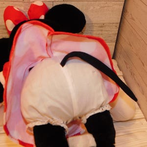 Op de afbeelding: Een pluche Minnie Mouse-pop, van achteren gezien, met witte bloomers met kanten rand en een rood-wit gestippelde jurk. Haar zwarte staart en oren zijn zichtbaar, samen met haar rode strik met witte stippen.