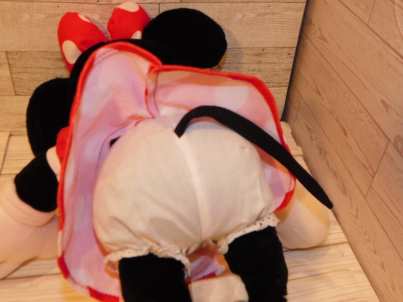 Op de afbeelding: Een pluche Minnie Mouse-pop, van achteren gezien, met witte bloomers met kanten rand en een rood-wit gestippelde jurk. Haar zwarte staart en oren zijn zichtbaar, samen met haar rode strik met witte stippen.