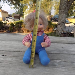 Puede incluir: Una mu&ntilde;eca vintage con cabello rubio que lleva una camisa a cuadros rojos y blancos y un mono azul. La mu&ntilde;eca est&aacute; sentada sobre una superficie de madera con una cinta m&eacute;trica a su lado.