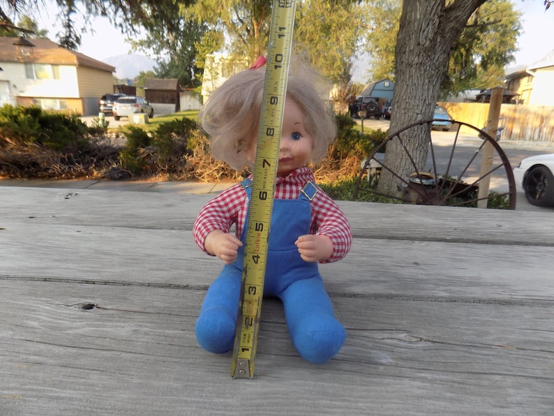 Puede incluir: Una mu&ntilde;eca vintage con cabello rubio que lleva una camisa a cuadros rojos y blancos y un mono azul. La mu&ntilde;eca est&aacute; sentada sobre una superficie de madera con una cinta m&eacute;trica a su lado.