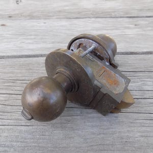 May include: A vintage brass door knob set featuring a spherical handle and a rectangular latch mechanism. The metal surface shows signs of age and oxidation, with a visible mounting plate and a small metal pin protruding from the side assembly.