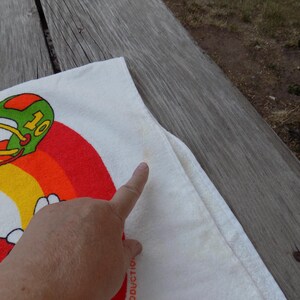Peut inclure: Serviette blanche avec un personnage de dessin anim&eacute; en uniforme de football, portant le num&eacute;ro 10. Le motif comprend un arc-en-ciel et un casque vert et orange. La serviette est sur une surface en bois.