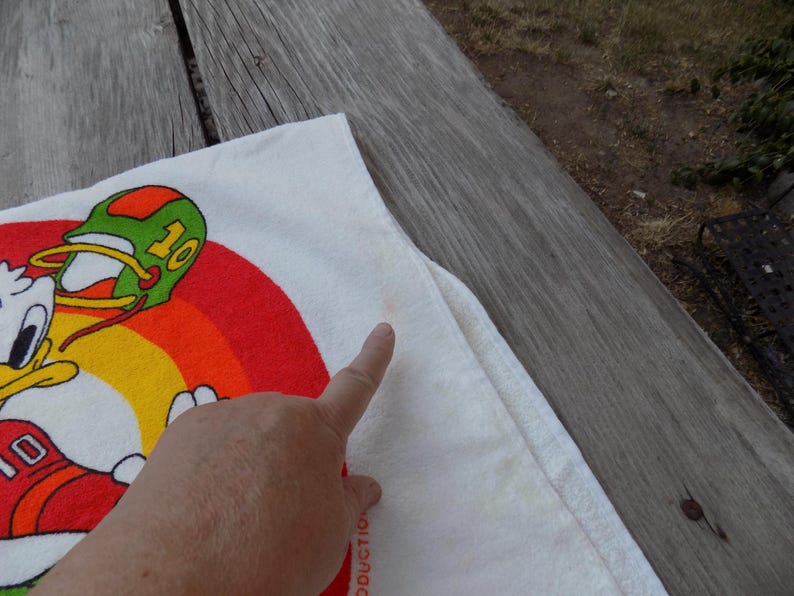 Peut inclure: Serviette blanche avec un personnage de dessin anim&eacute; en uniforme de football, portant le num&eacute;ro 10. Le motif comprend un arc-en-ciel et un casque vert et orange. La serviette est sur une surface en bois.