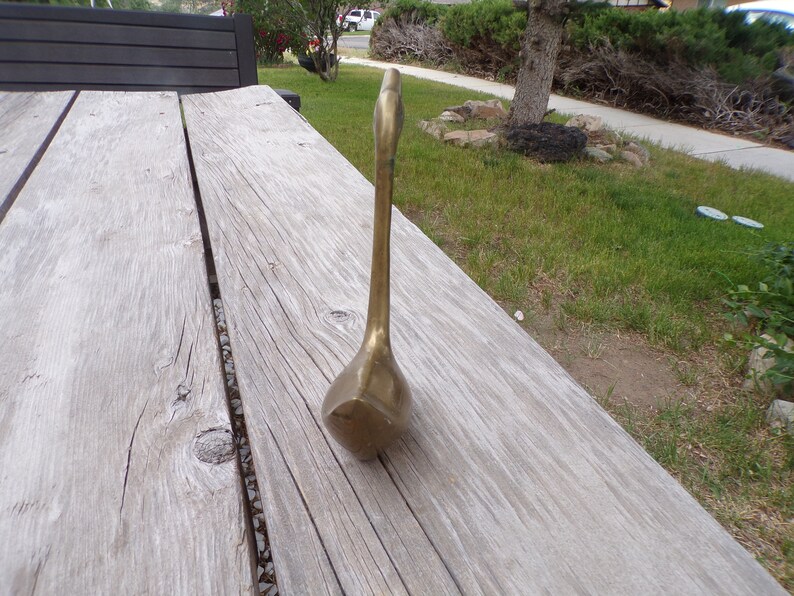 Pu&ograve; includere: Una statuetta di cigno in metallo dorato con un collo lungo e un corpo arrotondato. Il cigno &egrave; in piedi su una superficie di legno.