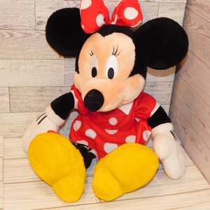 Op de afbeelding: Een pluche Minnie Mouse pop, met een rode jurk met witte stippen, een bijpassende strik en grote zwarte oren. De pop heeft gele voeten en witte handschoenen. Een klassiek Disney-personage.
