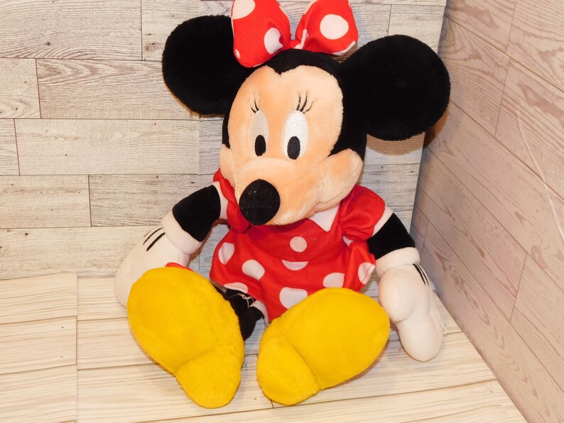 Op de afbeelding: Een pluche Minnie Mouse pop, met een rode jurk met witte stippen, een bijpassende strik en grote zwarte oren. De pop heeft gele voeten en witte handschoenen. Een klassiek Disney-personage.