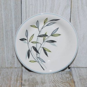 May include: A white ceramic bowl with a light blue rim and a botanical design. The design features black, yellow, and blue leaves and stems. The bowl is round and sits on a light-colored wooden surface.