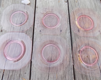 Può includere: Sei piatti vintage in vetro rosa con bordo smerlato e un anello rosa in rilievo al centro. I piatti sono quadrati e poggiano su una superficie di legno stagionato. I piatti sono di colore rosa tenue.