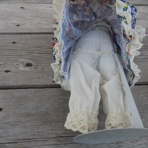 May include: A vintage doll with a floral patterned dress and white lace trim. The doll's white bloomers have lace trim at the ankles. The doll is positioned on a wooden surface.