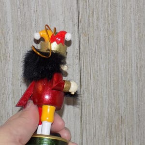 May include: A wooden nutcracker ornament with a red and gold costume, black fur trim, and a yellow and red jester hat. The nutcracker is holding a white object in its right hand.