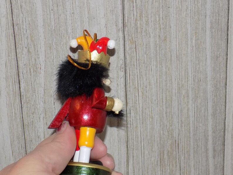 May include: A wooden nutcracker ornament with a red and gold costume, black fur trim, and a yellow and red jester hat. The nutcracker is holding a white object in its right hand.