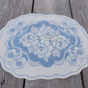 May include: A white and blue lace doily with a scalloped edge. The center features a floral design in white on a blue background. The doily is circular and rests on a wooden surface.