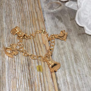 May include: A gold charm bracelet with four charms: a rocking horse, a bell, a carriage, and a shoe. The bracelet is shown on a weathered wooden surface, with white lace in the background.