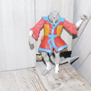 May include: A gray anthropomorphic rat action figure wearing a red and yellow robe with blue trim. The figure has white bandages on its arms and legs. The figure is posed on a wooden surface.