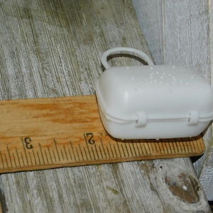 Op de afbeelding: Een kleine, witte plastic doos met een handvat en een scharnierende sluiting, ongeveer 7.6 cm lang, ligt op een houten liniaal. De doos heeft een gestructureerd oppervlak en is ontworpen om kleine items in te bewaren.