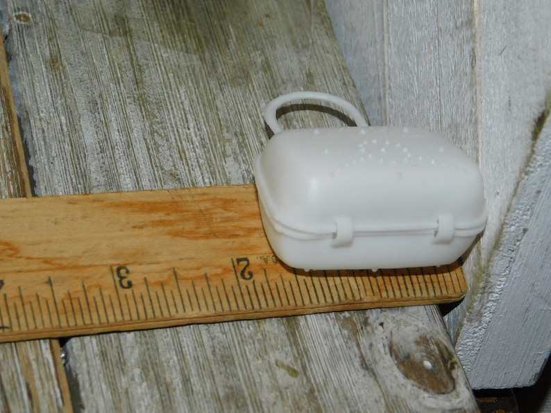 Op de afbeelding: Een kleine, witte plastic doos met een handvat en een scharnierende sluiting, ongeveer 7.6 cm lang, ligt op een houten liniaal. De doos heeft een gestructureerd oppervlak en is ontworpen om kleine items in te bewaren.