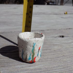 May include: A small, white ceramic cup with a speckled glaze and colorful abstract designs in shades of blue, green, and red.