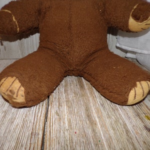May include: A brown teddy bear with worn fur and stitched details on the paws. The bear is sitting on a wooden surface.