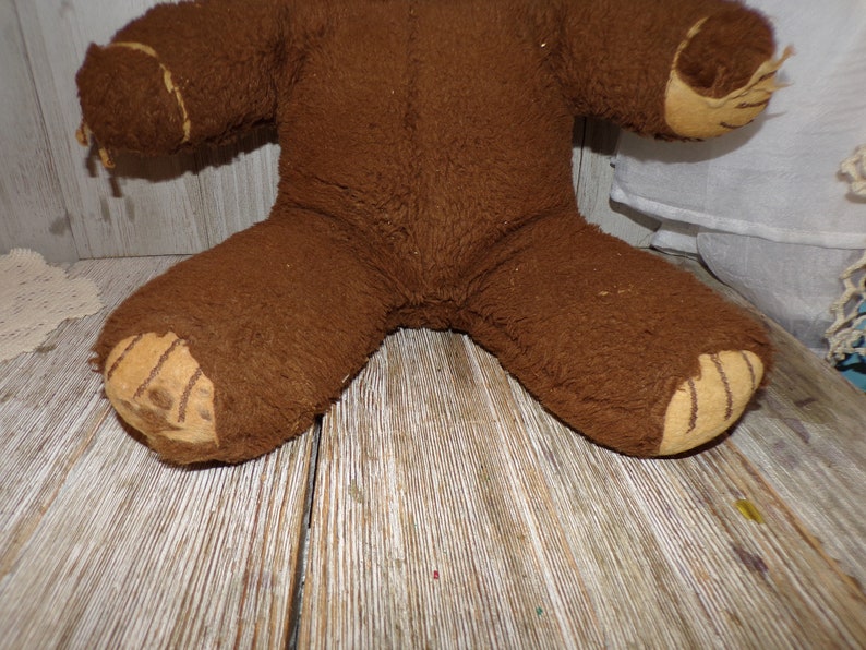 May include: A brown teddy bear with worn fur and stitched details on the paws. The bear is sitting on a wooden surface.