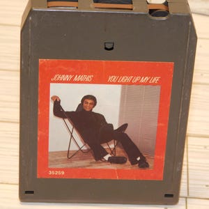 May include: An 8-track tape cartridge featuring Johnny Mathis' album "You Light Up My Life." The album art shows Mathis in a black outfit, seated in a chair. The cartridge is dark gray with a red label.