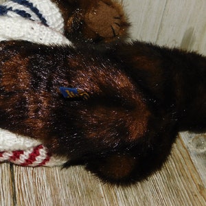 May include: Brown plush bear wearing a white, red and blue striped knitted sweater. The bear has a brown nose and black eyes. The sweater has a blue and white striped collar.