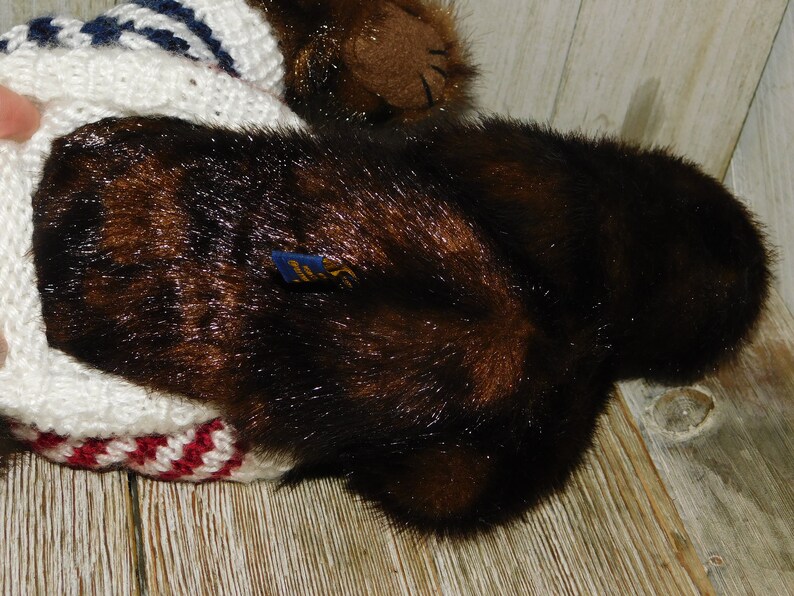 May include: Brown plush bear wearing a white, red and blue striped knitted sweater. The bear has a brown nose and black eyes. The sweater has a blue and white striped collar.