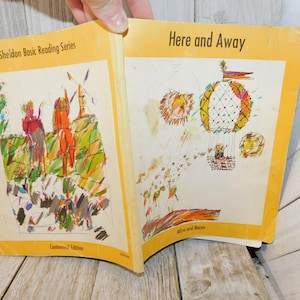 May include: Open book with colorful illustrations. The left page features figures near a windmill, while the right page shows hot air balloons and whimsical characters. Text reads "Here and Away" and "Sheldon Basic Reading Series."