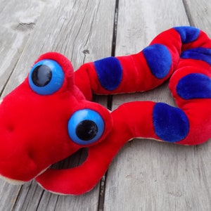 May include: A red and blue plush snake toy with large blue eyes. The snake is curled up on a wooden surface.