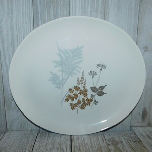 May include: A round, off-white ceramic plate with a botanical design. The design features a fern, and other plants in shades of blue, brown, and gray. The plate is set against a light wood background.