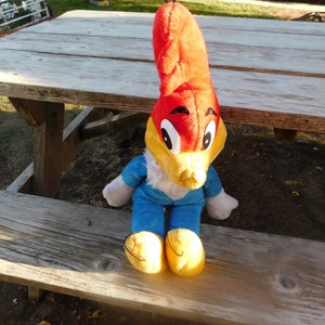 May include: A plush toy of Woody Woodpecker sitting on a wooden picnic table. The toy is red, yellow, and blue with a large beak and big eyes.