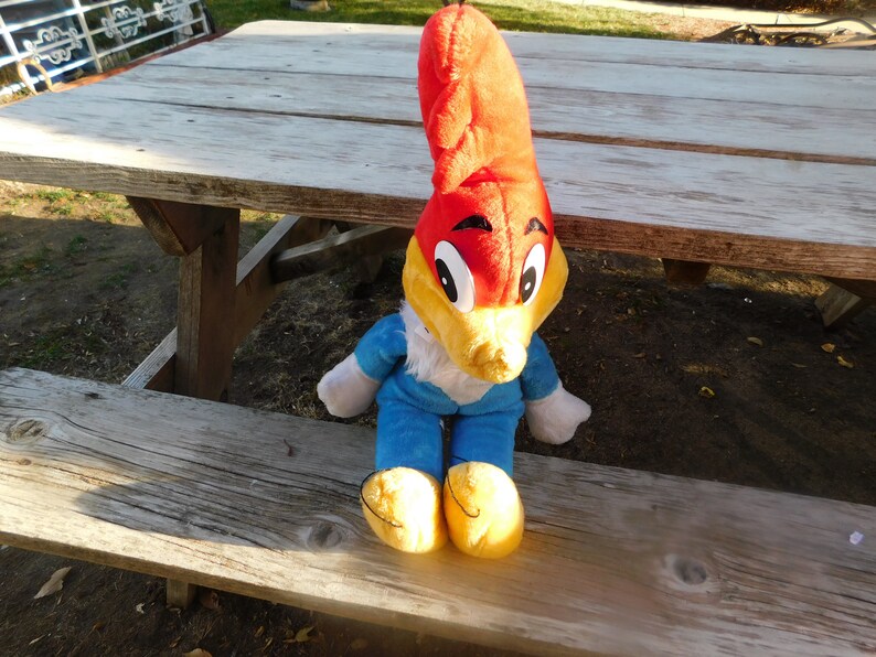 May include: A plush toy of Woody Woodpecker sitting on a wooden picnic table. The toy is red, yellow, and blue with a large beak and big eyes.