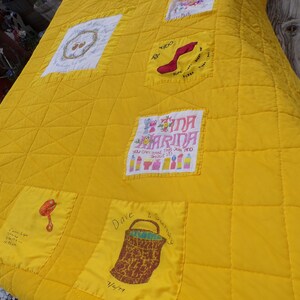 May include: A handmade yellow quilt with embroidered squares. The quilt features various embroidered designs, including a red shoe, a basket, and text that reads "Tina Marina". The quilt is quilted in a grid pattern.