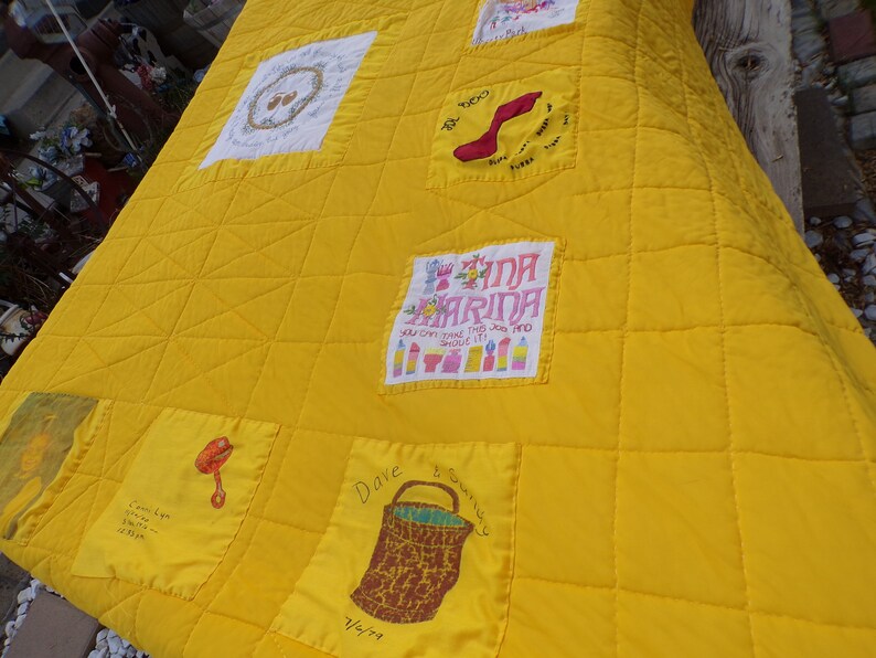 May include: A handmade yellow quilt with embroidered squares. The quilt features various embroidered designs, including a red shoe, a basket, and text that reads "Tina Marina". The quilt is quilted in a grid pattern.