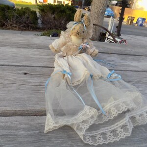 May include: A plush rabbit doll with tan fur wearing a cream-colored lace dress. The dress features a high collar, long sleeves, and a long, layered tulle skirt with scalloped lace trim. Light blue ribbons are tied at the waist and skirt.