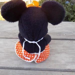 May include: A plush black mouse doll with large round ears and a yellow bow. The doll is wearing an orange and white polka dot skirt. The doll is sitting on a wooden surface.