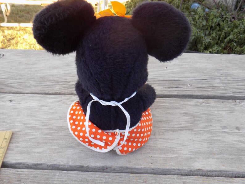 May include: A plush black mouse doll with large round ears and a yellow bow. The doll is wearing an orange and white polka dot skirt. The doll is sitting on a wooden surface.