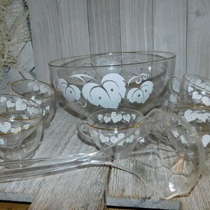 May include: A clear glass punch bowl set with a white leaf design. The set includes a large bowl, several cups with gold rims, and a ladle. The punch bowl and cups are arranged on a wooden surface.