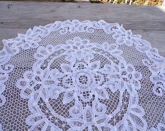May include: White crocheted lace tablecloth with a delicate floral pattern. The lace is intricate and detailed, with a circular design in the center.