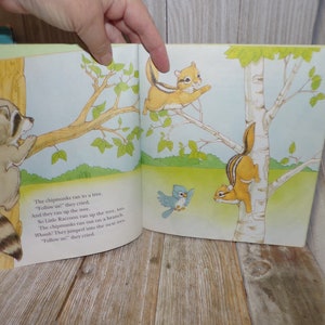 May include: A children's book illustration featuring a raccoon climbing a tree branch, two chipmunks climbing a birch tree, and a bluebird flying in the air. The text reads: "The chipmunks ran to a tree. "Follow us" they cried. And they ran up the tree. So Little Raccoon ran up the tree, too. The chipmunks ran out on a branch. Whee! They jumped into the next tree. "Follow us" they cried."