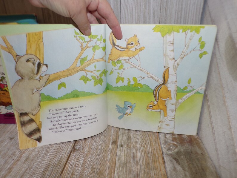 May include: A children's book illustration featuring a raccoon climbing a tree branch, two chipmunks climbing a birch tree, and a bluebird flying in the air. The text reads: "The chipmunks ran to a tree. "Follow us" they cried. And they ran up the tree. So Little Raccoon ran up the tree, too. The chipmunks ran out on a branch. Whee! They jumped into the next tree. "Follow us" they cried."