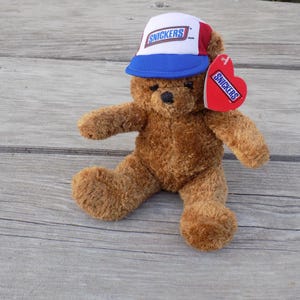 May include: A brown teddy bear wearing a white, blue, and red baseball cap with the "Snickers" logo. The bear has a red heart-shaped tag with the "Snickers" logo. The plush toy is sitting on a wooden surface.