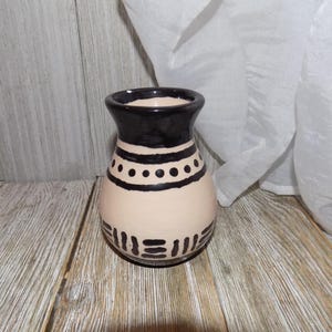 May include: A small, cream-colored ceramic vase with black geometric designs. The vase has a rounded body and a narrow opening.