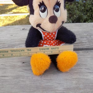 May include: A vintage Minnie Mouse plush toy with black fur, a red and white polka dot dress, and orange feet. A wooden ruler with the text "PBL NATIONAL PRESIDENT" is placed near the toy. The toy is sitting on a weathered wooden surface.