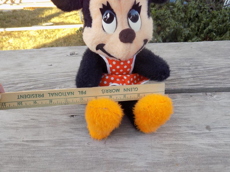 May include: A vintage Minnie Mouse plush toy with black fur, a red and white polka dot dress, and orange feet. A wooden ruler with the text "PBL NATIONAL PRESIDENT" is placed near the toy. The toy is sitting on a weathered wooden surface.