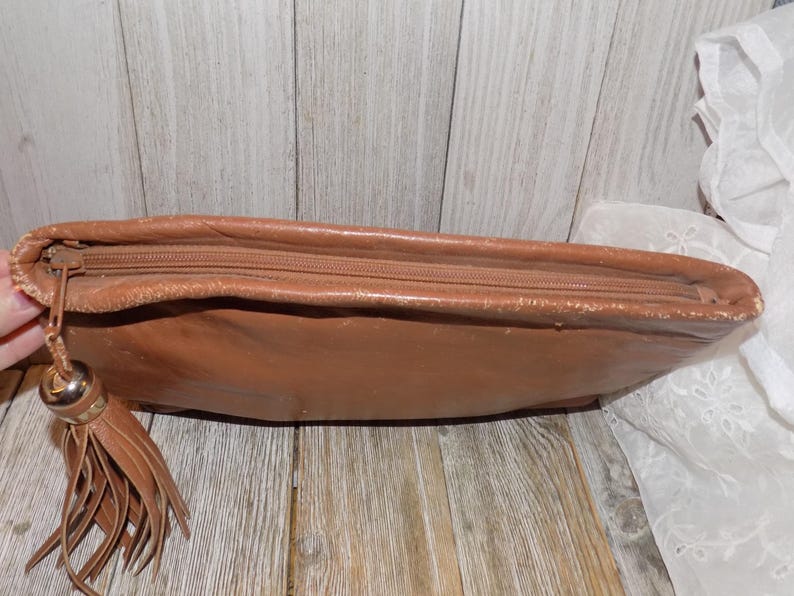 May include: A brown leather clutch bag with a zipper closure and a tassel. The bag has a worn look and is made of a soft, supple leather.