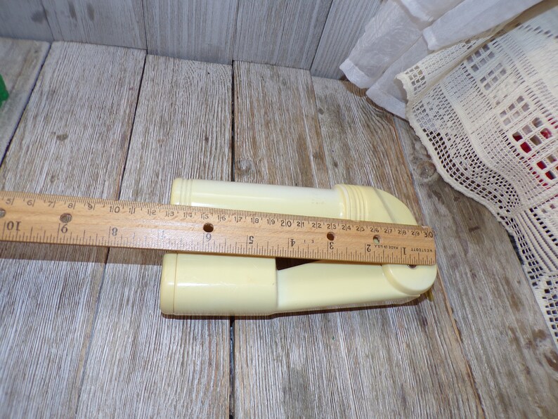 May include: A white plastic garlic press with a handle and a rotating mechanism. The press is about 7 inches long and is sitting on a wooden surface.