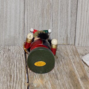 May include: A wooden nutcracker ornament with a red, blue, and green costume. The nutcracker has a white beard and a black hat with a red and white pom-pom. The nutcracker is holding a white ball in each hand.