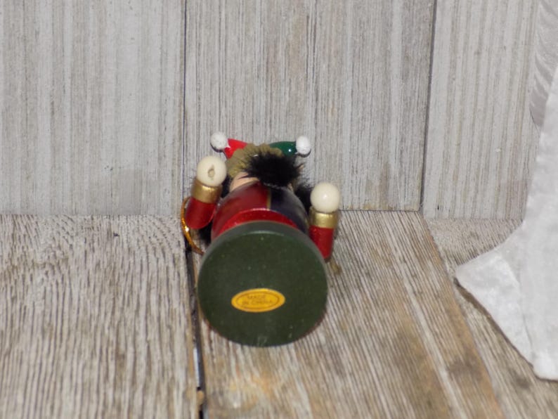 May include: A wooden nutcracker ornament with a red, blue, and green costume. The nutcracker has a white beard and a black hat with a red and white pom-pom. The nutcracker is holding a white ball in each hand.