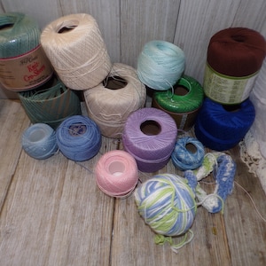 May include: A collection of yarn balls in various colors, including green, blue, purple, pink, brown, and white. Some of the yarn balls are in their original packaging, while others are loose. The yarn is arranged on a wooden surface.